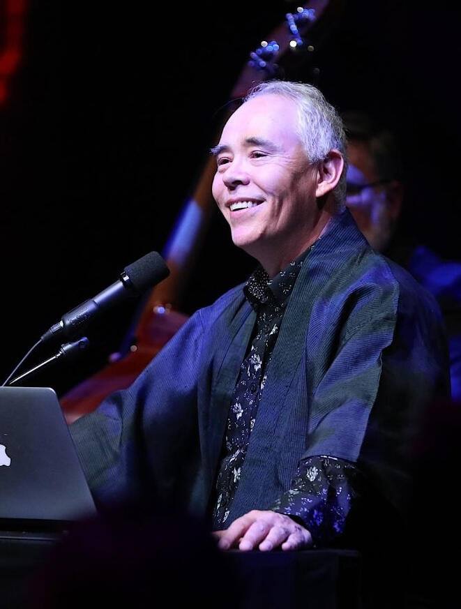 Tim Tamashiro smiling warmly while seated at a podium with a laptop, engaging with the audience during a live talk.